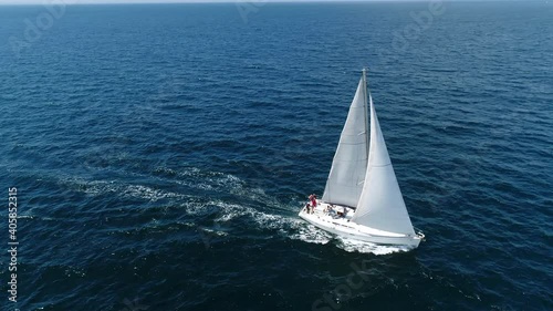 Aerial view. A white sailboat is moving fast on the waves of the sea. Flying above a boat in full sailing gear. Close hauled sailing in ocean. Yacht in windy and sunny day. Drone shot. photo