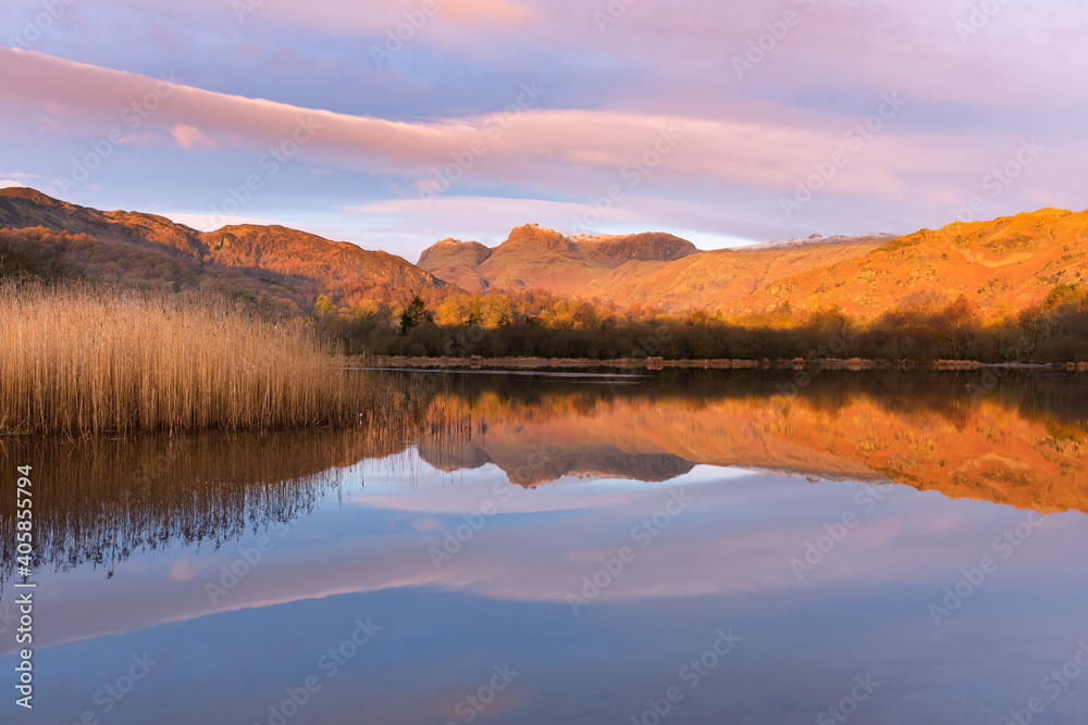 Mirrored reflections in calm lake with golden light from sunrise on mountain tops. Elterwater, Lake District, UK.