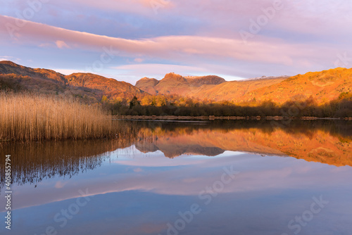 Mirrored reflections in calm lake with golden light from sunrise on mountain tops. Elterwater, Lake District, UK.