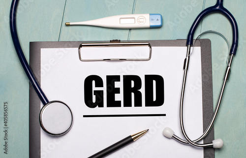 On a light blue wooden background, there is a paper labeled GERD, a stethoscope, an electronic thermometer, and a pen. Medical concept photo