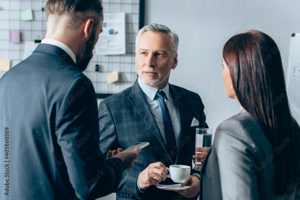 Mature advisor with cup of coffee looking at businessman with smartphone and businesswoman with glass of water on blurred foreground Mature advisor with cup of coffee looking at businessman with smartphone and businesswoman with glass of water on blurred foreground