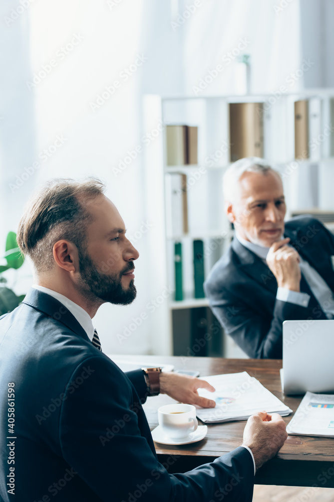 Businessman sitting near cup of coffee, papers and advisor on blurred background Businessman sitting near cup of coffee, papers and advisor on blurred background