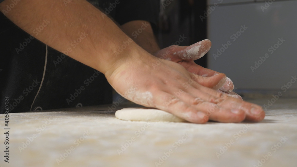Close up arms of cook forming pastry on a wooden surface at cuisine. Male hands of chef shaping floured dough for pizza on a kitchen table at restaurant. Concept of cooking food. Slow motion