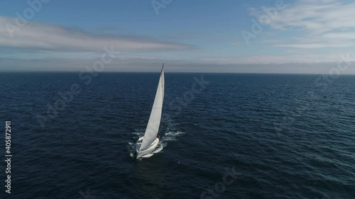Aerial view. A white sailboat is moving fast on the waves of the sea. A boat in full sailing gear floats on camera. Close hauled sailing in ocean. Yacht in windy and sunny day. Drone shot.