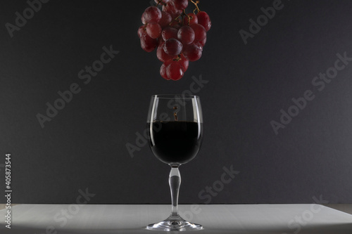 Copa de cristal con uvas goteando vino en ella. Goteo de vino en copa de cristal sobre fondo negro y superficie blanca. Iluminación lateral.