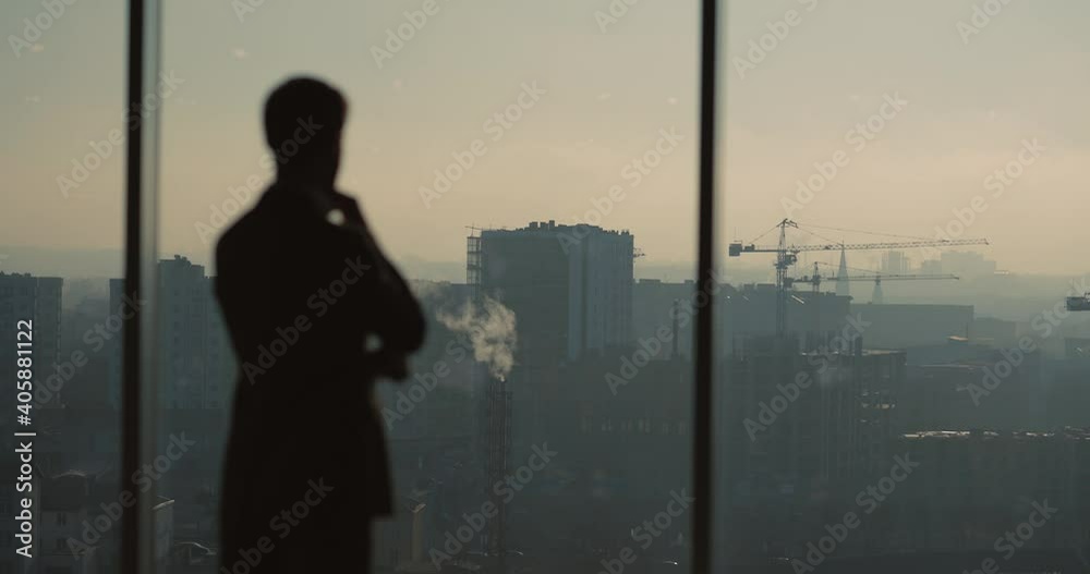Silhouette of successful businessman contemplating business idea looking through large window at city buildings. Modern cityscape. Sunsets.