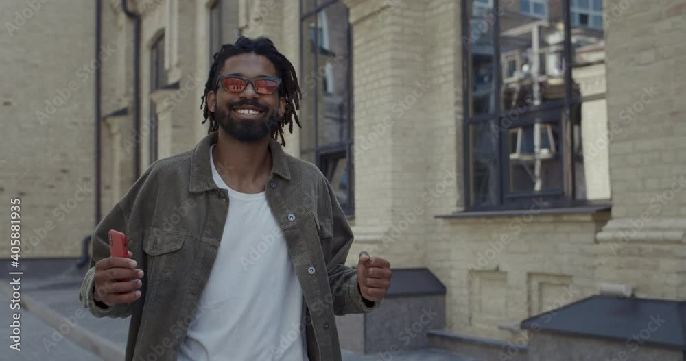 Joyful guy with dreadlocks smiling and dancing while looking to camera outdoors. Bearded afro american man in stylish red glasses having good mood and singing while walking at street Joyful guy with dreadlocks smiling and dancing while looking to camera outdoors. Bearded afro american man in stylish red glasses having good mood and singing while walking at street