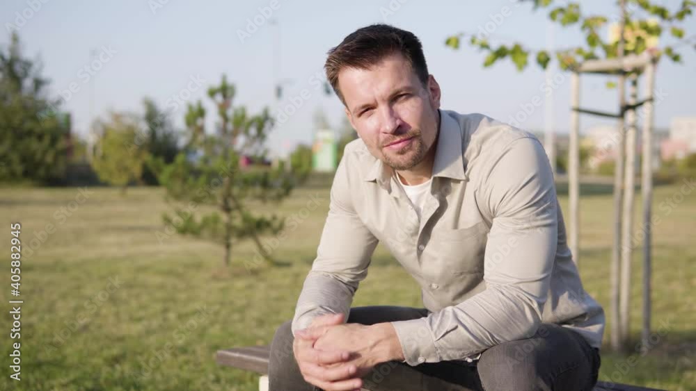 A young Caucasian man smiles at the camera as he sits in a park in an urban area on a sunny day - buildings in the blurry background A young Caucasian man smiles at the camera as he sits in a park in an urban area on a sunny day - buildings in the blurry background