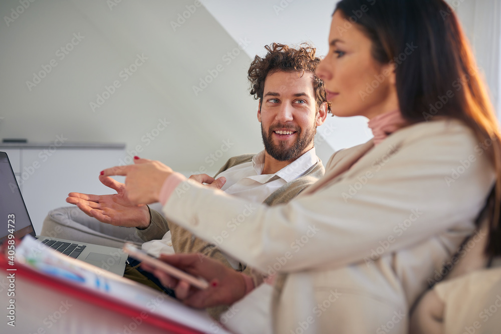 Fototapeta premium Cheerful business couple sitting and discussing on the couch in a an apartment. Business, couple, leisure, together
