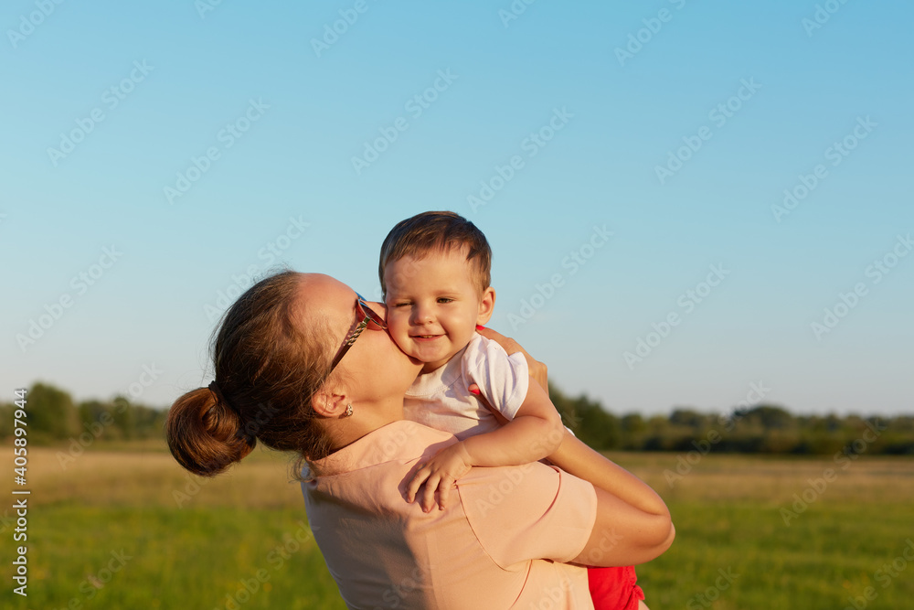 Mother holding her little child in hands while walking outside on beautiful nature, mommy kissing her toddler on cheek, family expressing happiness, cute small girl with mom. Mother holding her little child in hands while walking outside on beautiful nature, mommy kissing her toddler on cheek, family expressing happiness, cute small girl with mom.