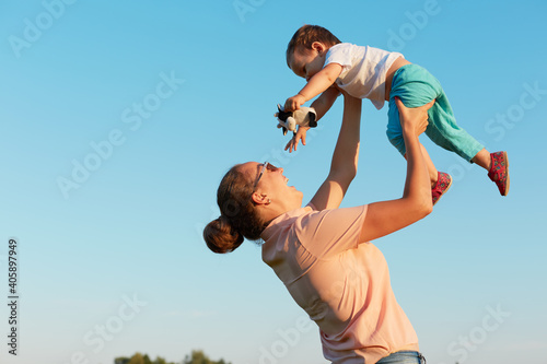 Happy young mother with toddler in hands, spending time isolated over blue sky background, lady with hair bun spending time with her little child flying .