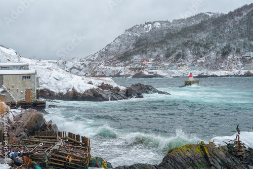 A winter's scene of St. John's harbor, Newfoundland. The sky is overcast. There's a lighthouse at the land's point along with small homes.  The bottom part of the picture has a marine marker   photo