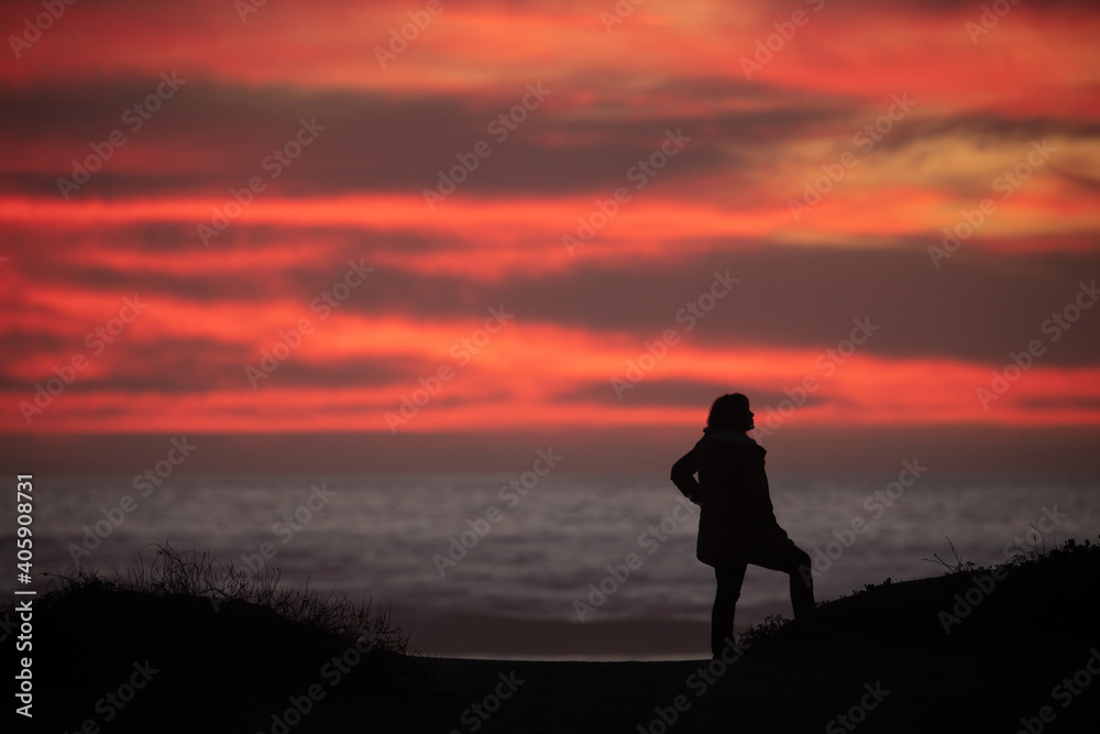 Silhouette of woman standing on hill overlooking the ocean with a dramatic orange sunset 
