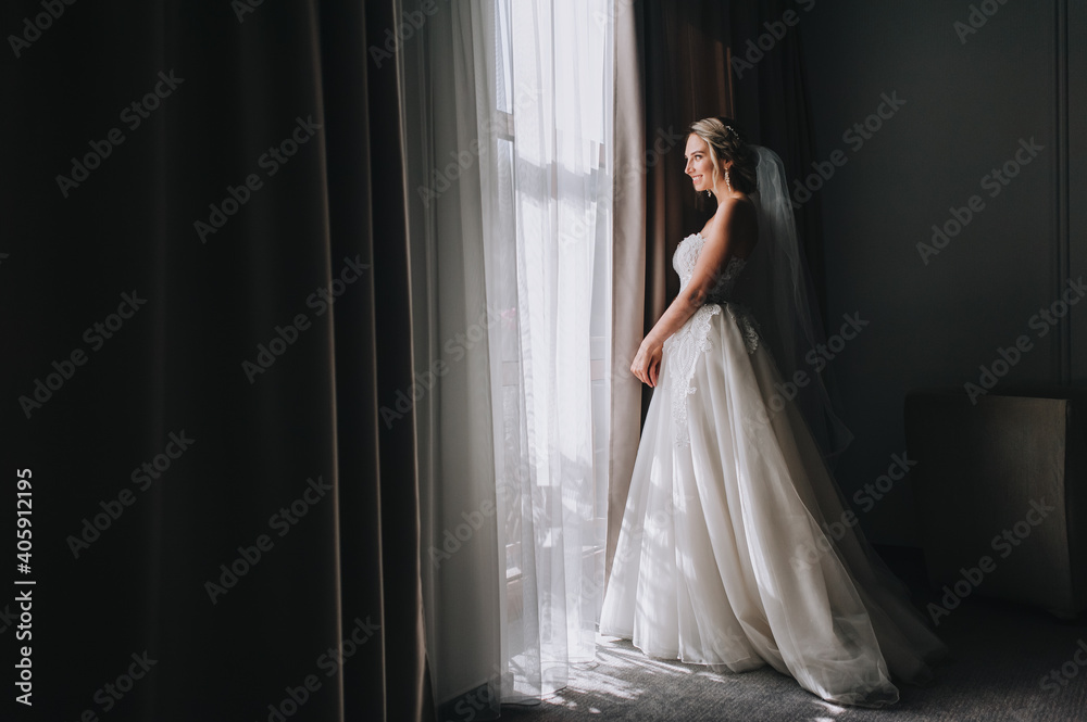 A beautiful blonde bride in a white long dress stands near the window with curtains in the room of a posh hotel. Wedding portrait. A beautiful blonde bride in a white long dress stands near the window with curtains in the room of a posh hotel. Wedding portrait.