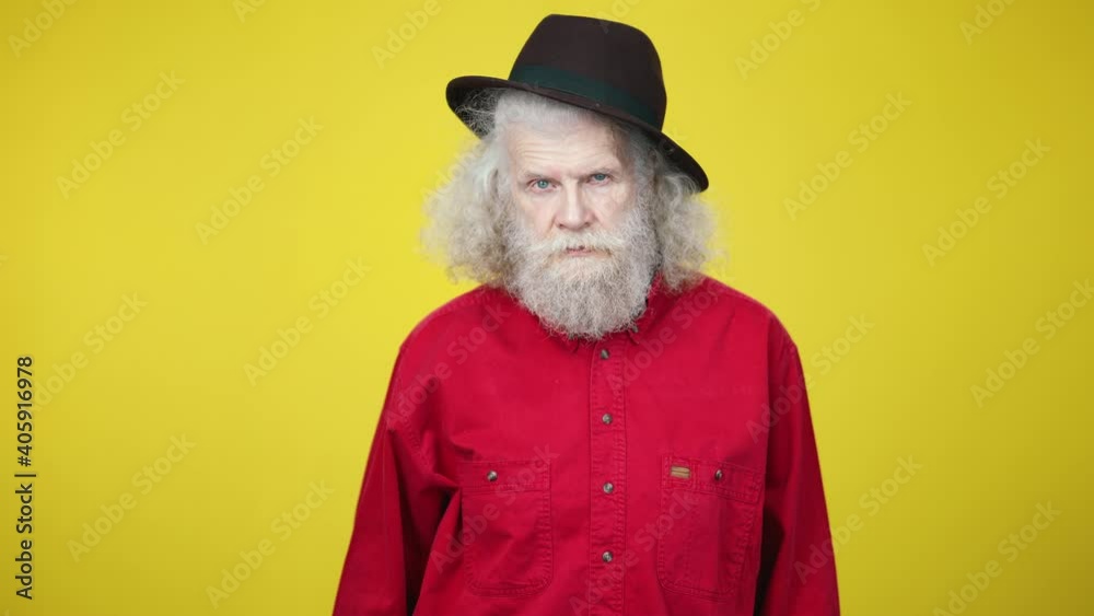 Portrait of elegant senior man in hat taking off sunglasses and winking with serious facial expression. Handsome grey-haired bearded Caucasian male retiree posing at yellow background. Elegance Portrait of elegant senior man in hat taking off sunglasses and winking with serious facial expression. Handsome grey-haired bearded Caucasian male retiree posing at yellow background. Elegance