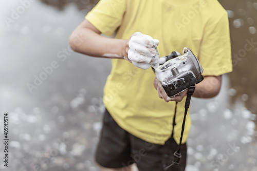 A boy in a yellow t-shirt and shorts is standing near the water in nature and washing a camera with soap and foam. The concept child is a bully. photographers day photo