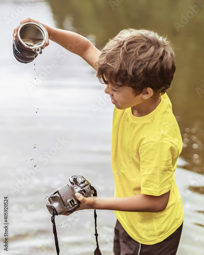 A boy stands in the river on outdooor and destroys a mirrorless photo camera. The concept of obsolescence of technology. Photographers day, humor. Toned picture. Soft light. Instagram photo.