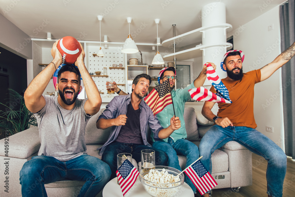 Excited male friends cheering while watching American football match on sofa at home.