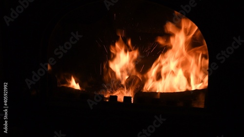 Blurred fiery background with spark traces and copy space. Rustic fireplace with burning firewood and glowing flame inside with bright sparks. Cozy warm light in furnace in night darkness