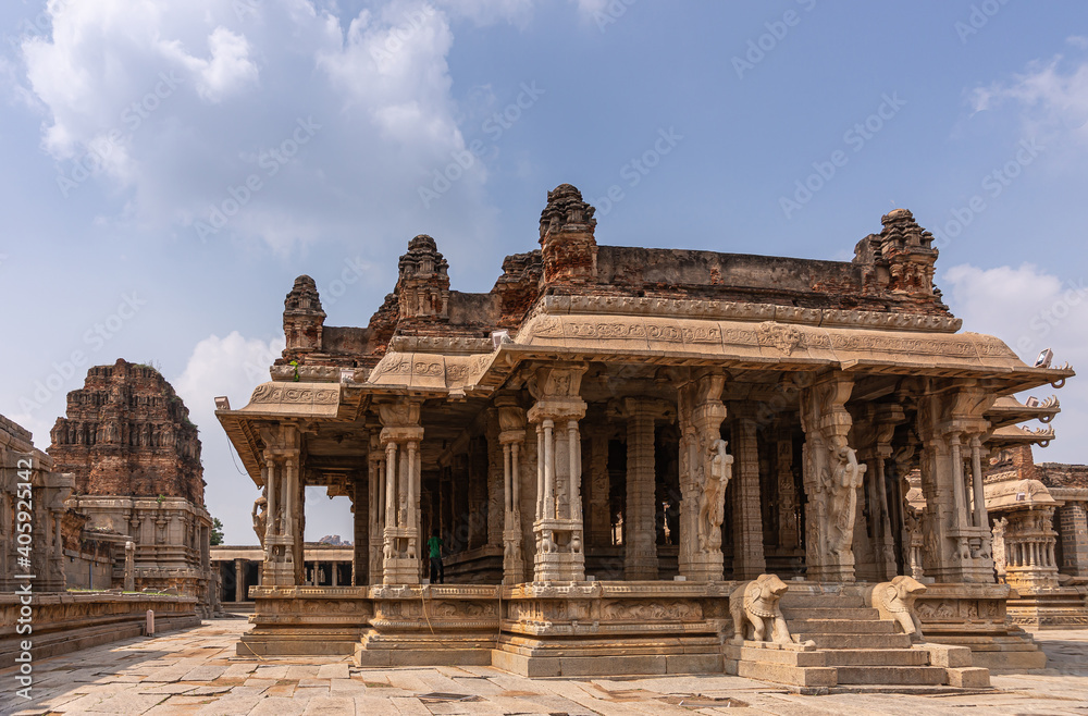 Hampi, Karnataka, India - November 5, 2013: Vijaya Vitthala Temple. Group of brown stone sanctuaaries with or without towers and decorated columns under blue cloudscape.