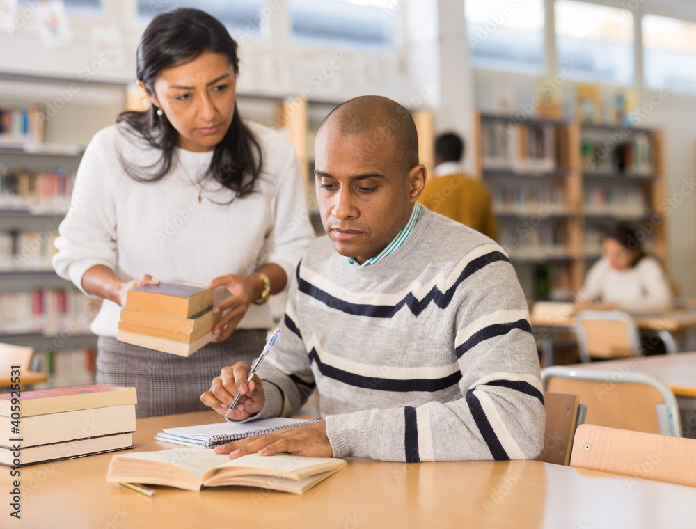 Fototapeta premium Female librarian helping focused adult hispanic student preparing to exam, bringing him books