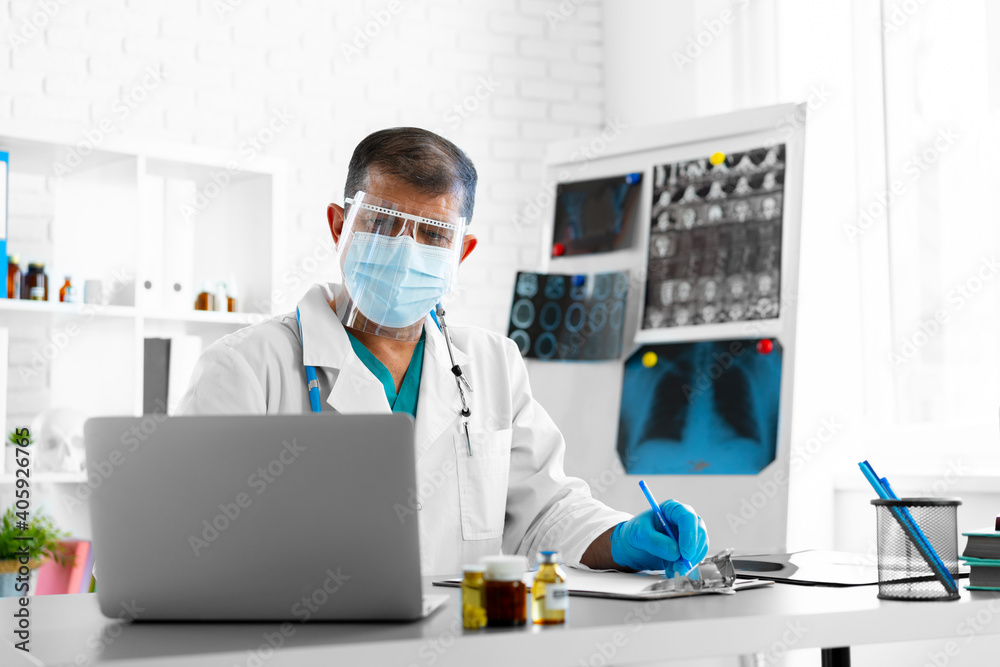 Middle-aged male doctor in face shield and mask sitting at the table at his office Middle-aged male doctor in face shield and mask sitting at the table at his office