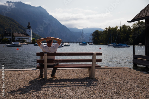 The middle aged man relaxing by lake, watching at water and mountains.