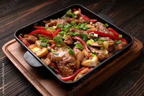 Fried meat with vegetables, green onions, bell peppers and mushrooms, in a frying pan, on a wooden background
