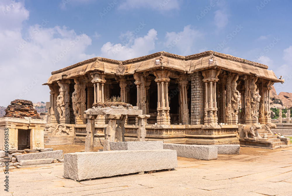 Hampi, Karnataka, India - November 5, 2013: Vijaya Vitthala Temple. Smaller side brown stone mandapam hall with elephant steps and sculpted pillars under blue cloudscape.