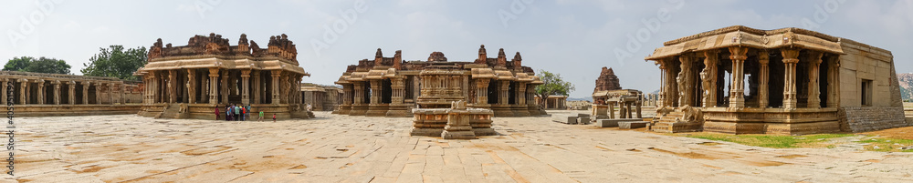 Hampi, Karnataka, India - November 5, 2013: Vijaya Vitthala Temple. Panorama shot of main brown stone buildings under light blue sky and beige stone courtyard uip front. People add color.