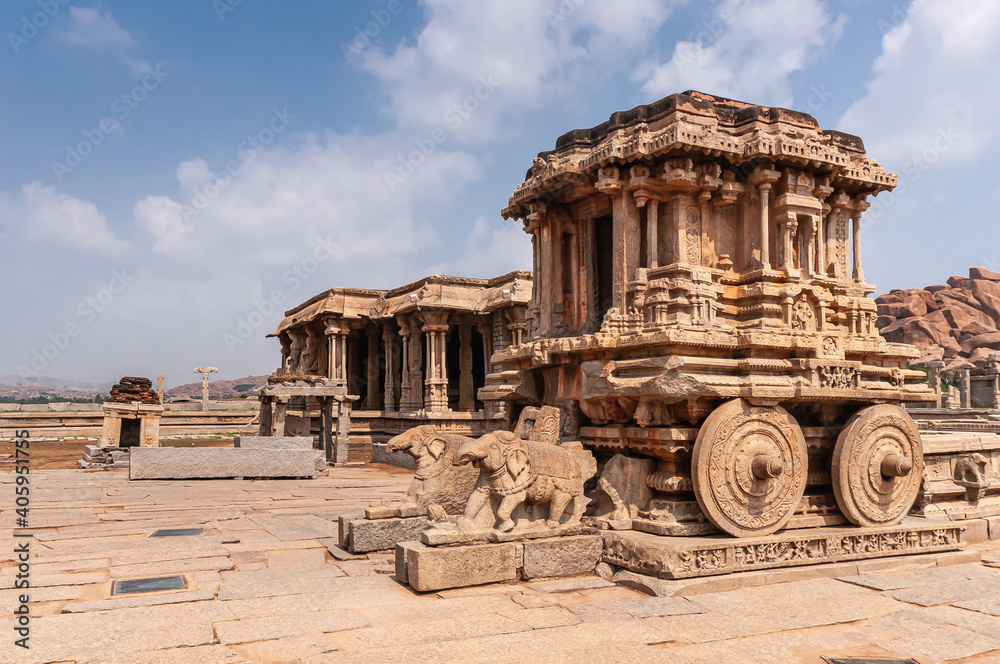 Hampi, Karnataka, India - November 5, 2013: Vijaya Vitthala Temple. Front and side of brown stone chariot under blue cloudscape.