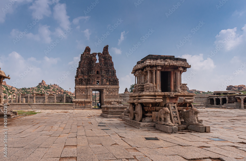 Hampi, Karnataka, India - November 5, 2013: Vijaya Vitthala Temple. Wide view on brown stone chariot from the front under blue cloudscape with red stone east Gopuram in back.