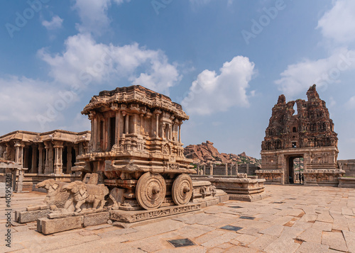 Hampi, Karnataka, India - November 5, 2013: Vijaya Vitthala Temple. Closeup of brown stone chariot from the front under blue cloudscape with red stone east Gopuram in back.