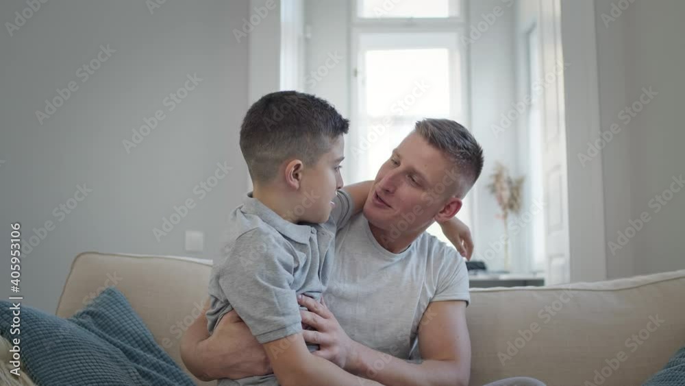 Young Man Sitting On Sofa In Apartment. His Son Runs To Him. Young Man Sitting On Sofa In Apartment. His Son Runs To Him.