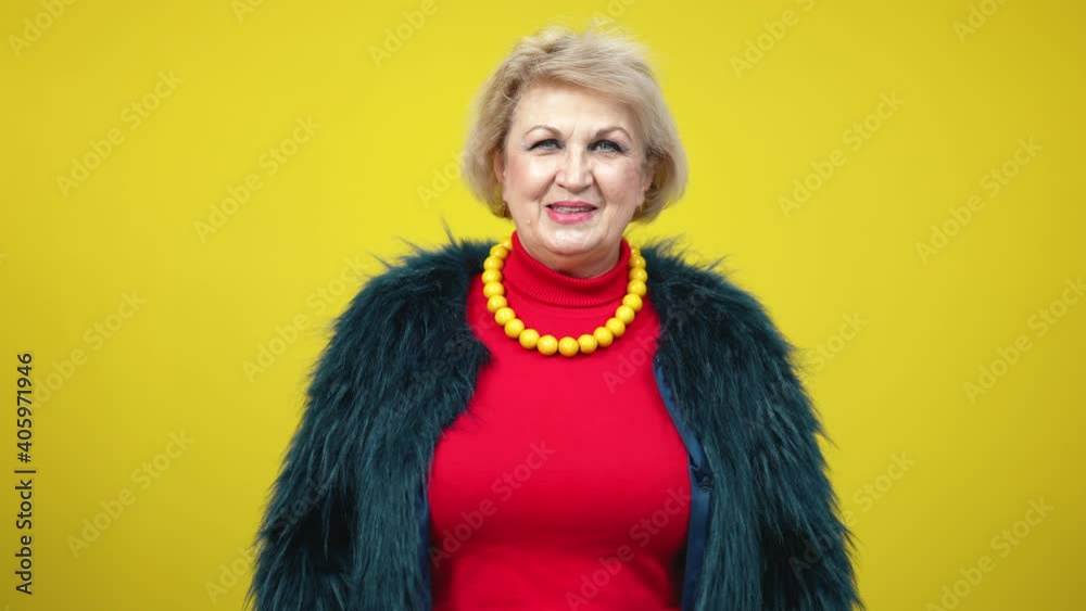 Elegant confident senior woman showing thumb up and winking on yellow background. Portrait of stylish Caucasian lady in fur coat posing looking at camera smiling.