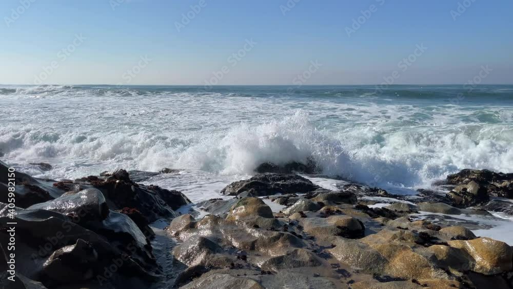 Rough, choppy white waves splashing over rocks on the beach on a bright sunny day with blue sky. Slow motion. Rough, choppy white waves splashing over rocks on the beach on a bright sunny day with blue sky. Slow motion.