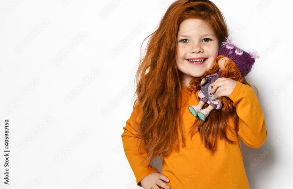 Portrait of frolic smiling five-year-old red-haired kid girl in orange sweatshirt holding small redhair doll in hands on her shoulder over white background with copy space