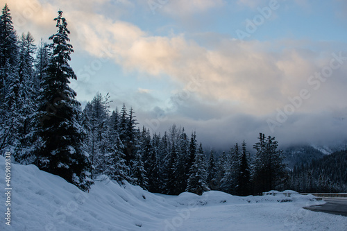 Winter highway among snow-covered mountains and tall fir trees at sunset.
