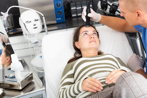Young woman getting facial electroporation procedure from experienced male cosmetologist in aesthetic medicine office