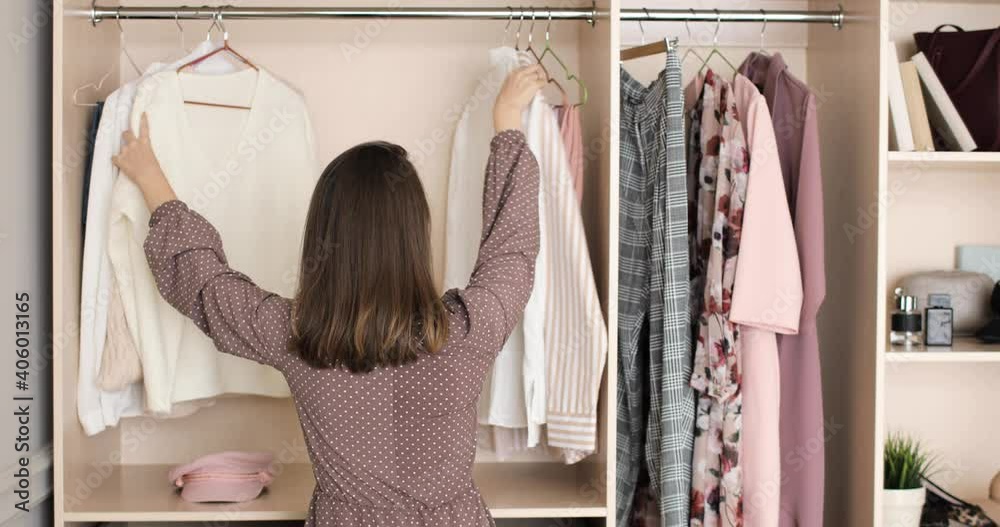 Female trying to picks up an outfit in home wardrobe. Young woman is choosing clothes on a racks in closet searching what to wear, back view. Concept of everyday dilemma in outfit choosing.