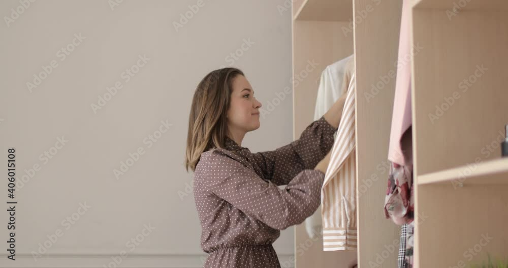 Young pretty woman is choosing clothes in wardrobe at home trying a white sweater looking at mirror, side view. Concept of everyday dilemma in outfit choosing. Big closet with clothes. Young pretty woman is choosing clothes in wardrobe at home trying a white sweater looking at mirror, side view. Concept of everyday dilemma in outfit choosing. Big closet with clothes.