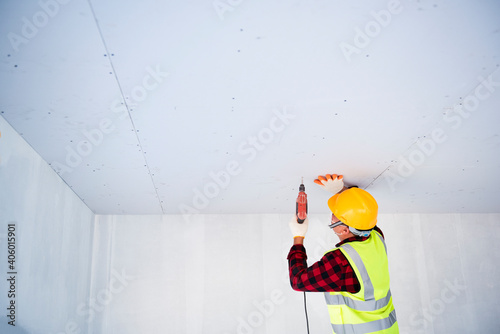 A young Asian construction worker in safety clothing and work gloves is fastening the drywall ceiling to the metal frame using an electric screwdriver on the ceiling covered in shiny aluminum foil.