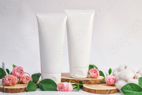 White unbranded tubes for cosmetics with wood cuts, small pink roses and cotton flowers on white background. Natural cosmetic product mock up, branding concept