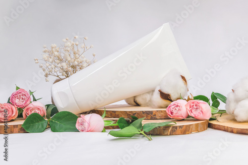White unbranded tube for cosmetics with wood cuts, small pink roses and cotton flowers on white background. Natural cosmetic product mock up, branding concept