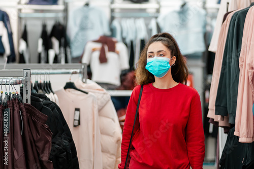 Consumerism. Portrait of young caucasian woman in a medical mask choosing clothes in a store. The concept of shopping during a viral pandemic