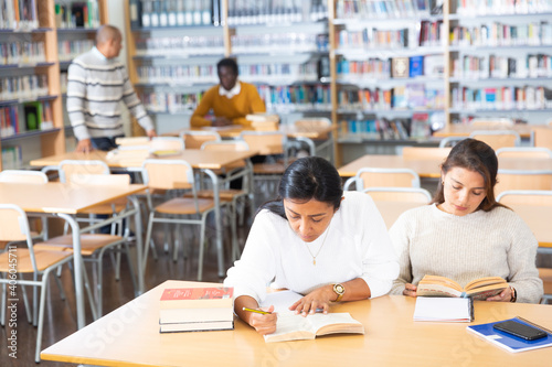 Positive young women preparing to exam in library, reading books and writing in notepads