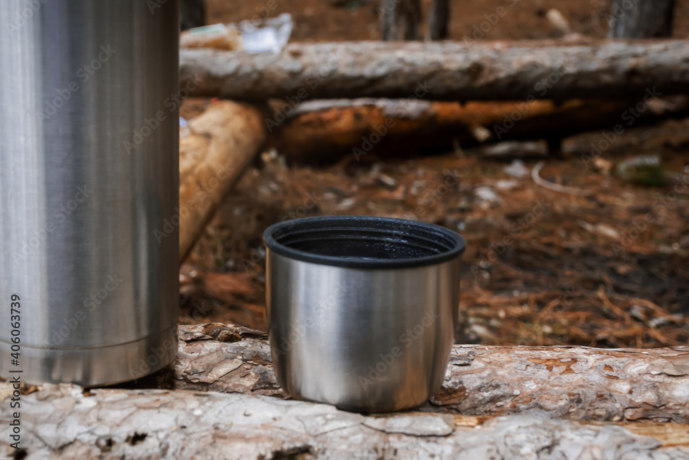 Thermos of tea in the forest close-up. Metal thermos with a mug in selective focus. One-day hike in winter in the mountains and forest.A place to relax.The concept of hiking, tourism, active lifestyle