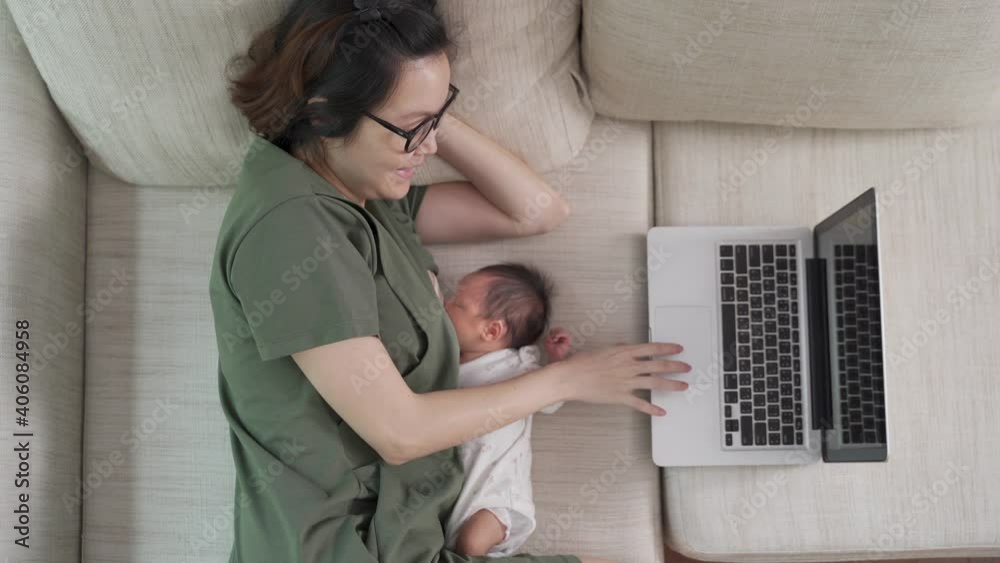 asian woman mother working from home on computer laptop and feeding breast milk to baby infant. parent multitasking on sofa couch in living room.