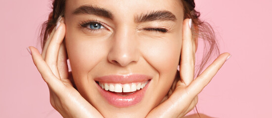Skincare and beauty. Close up of smiling young woman winking at camera, touching shiny healthy skin, nourished face after tea tree and lemon facial serum and moisturizer, pink background