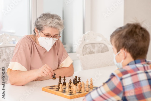 Senior grandmother and her grandson wearing protective face masks play chess at home during quarantine Coronavirus (Covid-19) epidemic